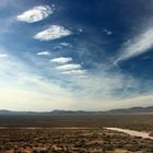 Weite Wüstenlandschaft mit Bergen am Horizont, darüber ein Himmel mit vereinzelten Wolken, unten fährt ein rotes Auto auf einem Weg.