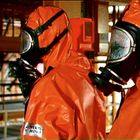 Zwei Menschen in orangen Schutzanzügen mit Gasmasken in industrieller Umgebung; Text: "PERIT NOVA".