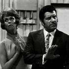 Eine Frau und ein Mann stehen nebeneinander. Die Frau hat kurz geschnittenes Haar, der Mann trägt einen Anzug mit gemusterter Krawatte. Sie wirken überrascht oder besorgt.