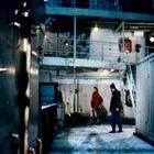 Zwei Personen stehen auf einem Schiffsdeck. Die Umgebung wirkt industriell, mit Containern und Metallstrukturen.