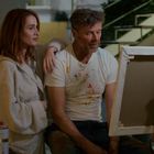 Zwei Personen, eine Frau im Bademantel und ein Mann mit Farbflecken auf dem T-Shirt, betrachten zusammen eine Leinwand.