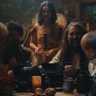 Eine Gruppe fröhlicher Menschen sitzt an einem Tisch, dazu eine Frau im Hintergrund. Es herrscht eine gesellige Atmosphäre.