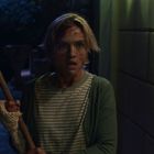 Eine Frau mit kurzem Haar, blutverschmiert, hält eine Axt und steht schockiert im Freien bei Nacht.