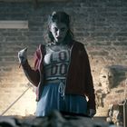 T-Shirt mit Text "LO DO". Frau in roter Jacke in einem Raum mit Backsteinwand, Masken und Seilen im Hintergrund.