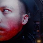 Ein Mann mit Nachdenklichkeit im Gesicht, beleuchtet von rotem Licht, in einem engen, technologischen Raum.