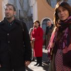 Ein Mann und eine Frau sind im Vordergrund, umgeben von mehreren Personen. Die Gebäude im Hintergrund wirken elegant.