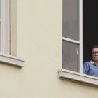 Eine Frau mit Brille schaut nachdenklich aus einem geöffneten Fenster eines gelben Gebäudes.