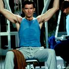 Zwei Männer in blauen Tanktops trainieren in einem Fitnessstudio an Kraftmaschinen. Der Fokus liegt auf dem vorderen Mann.
