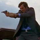 Ein Mann in einem Anzug zielt mit einer Pistole. Im Hintergrund ist ein Auto erkennbar.