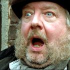 Ein älterer Mann mit blauen Augen und Bart hat einen erstaunten Gesichtsausdruck. Hintergrund ist eine Ziegelmauer.