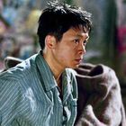 Ein Mensch mit kurzem dunklem Haar schaut ernst ausdruckslos in die Ferne. Die Person trägt ein gestreiftes Hemd.