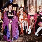 Sechs Frauen sitzen in farbenfroher Kleidung auf Stühlen, einige mit offenen Mündern, als ob sie singen oder überrascht sind.