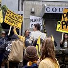Mehrere Personen halten Schilder mit der Aufschrift "PLANET PANIC". Sie stehen vor einem Gebäude im Regen.