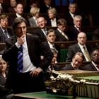 Ein Mann im Anzug steht nachdenklich inmitten einer Versammlung auf grünen Bänken in einem formellen Raum.