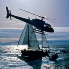 Segel mit „GREENPEACE“ Schriftzug. Ein Hubschrauber fliegt dicht über einem Segelboot im Meer bei Sonnenuntergang.