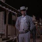 Ein Mann in einem Western-Outfit steht vor einem Holzgebäude, im Hintergrund sind zwei Pferde zu sehen, es ist Nacht.