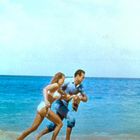 Ich kann den Text nicht identifizieren. Ein Mann und eine Frau laufen am Strand entlang, das Meer ist im Hintergrund sichtbar.