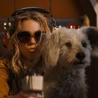 Ich kann den Text nicht erkennen. Eine Frau mit Kopfhörern und Sonnenbrille sitzt in einem Cockpit neben einem Hund.