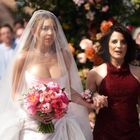 Zwei Frauen, eine im Hochzeitskleid mit Blumenstrauß, die andere in einem roten Kleid, Hand in Hand gehend. Hintergrund: Gäste.