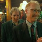 Zwei Männer in einem festlichen Raum, einer mit Brille und Schirmchendrink lächelt. Hinter ihnen viele Menschen und Dekorationen.