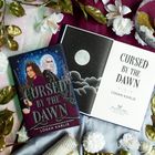 Zwei Bücher mit dem Titel "Cursed by the Dawn" von Logan Karlie, umgeben von Blumen und goldenen Blättern.