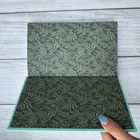 Ein geöffnetes Notizbuch zeigt Blättermuster auf beiden Seiten. Eine Hand mit blau lackierten Nägeln hält die Ecke.