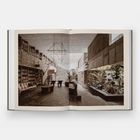 Ein offenes Buch zeigt eine Bibliothek mit hohen Regalen, einer modernen Skulptur, Pflanzen und einer Steintreppe.