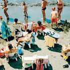 Menschen entspannen am Strand auf Plastikstühlen; Handtücher hängen, Kinder spielen am Wasser.