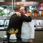 Zwei Köche umarmen sich in einer belebten Restaurantküche, umgeben von Kürbissen und Kräutern.