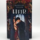 Der Text lautet: "Madeline Taylor HEIR". Davor ist eine Illustration eines Paares, das sich nahe steht, vor einem dekorativen Hintergrund.