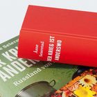 Rotes Buch mit Titel "DER KRIEG IST ANDERSWO" von Åsne Seierstad, auf einem weiteren Buch liegend.