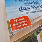 Roter Aufkleber: "SPIEGEL Bestseller-Autorin". Hintergrund zeigt einen Buchumschlag mit blauem Himmel.