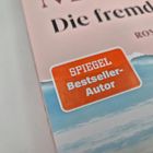 Text: "SPIEGEL Bestseller-Autor". Ein Buchcover mit dem Titel "Die fremde Frau" und blauem Himmel im Hintergrund.