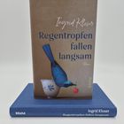 "Regentropfen fallen langsam" von Ingrid Kloser. Ein blauer Vogel pickt nach einer Kirsche, ein Buch liegt darunter.