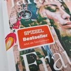 Rotes "SPIEGEL Bestseller"-Label, darunter "Jetzt als Taschenbuch". Hintergrund: Collage mit Frauengesicht und Rakete.