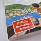 Auf einem Buchcover steht "SPIEGEL Bestseller-Autorin". Eine Frau im gelben Badeanzug sitzt in einem Boot.