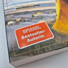 Text: "SPIEGEL Bestseller-Autorin." Ein orangefarbenes Logo auf einem Buchcover mit Dünen und einer Frau in gelbem Kleid.