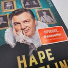 Text "SPIEGEL Bestseller Jetzt als Taschenbuch" in rotem Sticker. Person in Rüschenkleidung vor Ahnenbildern im Hintergrund.