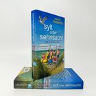 Aufrecht stehendes Buch "Sylt oder Sehnsucht" von Claudia Thesenfitz. Strandmotiv mit Menschen und Schwimmtieren.
