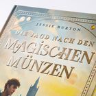 Text: "Jessie Burton: Die Jagd nach den Magischen Münzen". Buchcover mit Himmel und junger Mann im Profil.
