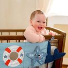 Ein lachendes Baby lehnt sich aus einem Holzbabybett. Daran hängen Stoffdekorationen: ein Rettungsring und ein Seestern.