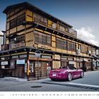 Eine traditionelle japanische Holzfassade vor der ein pinkfarbener Porsche 718 Boxster parkt.