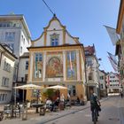 Text: "Weinstube Kinz". Historisches Gebäude mit Wandgemälde und Straßencafé, Radfahrer passierend. Klarer Himmel.