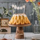 Eine Torte auf Holzständer, dekoriert mit Margariten-Zahnstocher, umgeben von Blumen, in rustikalem Ambiente.