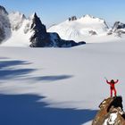 Ein Bergsteiger in roter Kleidung steht mit erhobenen Armen auf einem Felsen, umgeben von schneebedeckten Bergen und Gletschern.