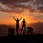 Ich kann diesen Text nicht identifizieren. Zwei Personen stehen bei Sonnenuntergang auf einem Tisch, daneben Fahrräder und Berge.