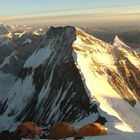 Keine erkennbaren Texte. Mehrere orangefarbene Zelte auf einem schneebedeckten Berggipfel bei Sonnenaufgang.