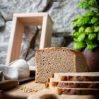 Ein Laib Vollkornbrot, teilweise geschnitten, auf einem Holzbrett. Im Hintergrund sind eine Steinmauer und grüne Kräuter.