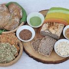 Verschiedene Brotsorten, darunter ein Laib in einem bunten Tuch. Umgeben von Schalen mit Leinsamen, Hafer und grünem Pulver.