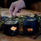 Eine Hand streut Kräuter über einem Stövchen mit brennendem Teelicht. Daneben trockene Pflanzen.